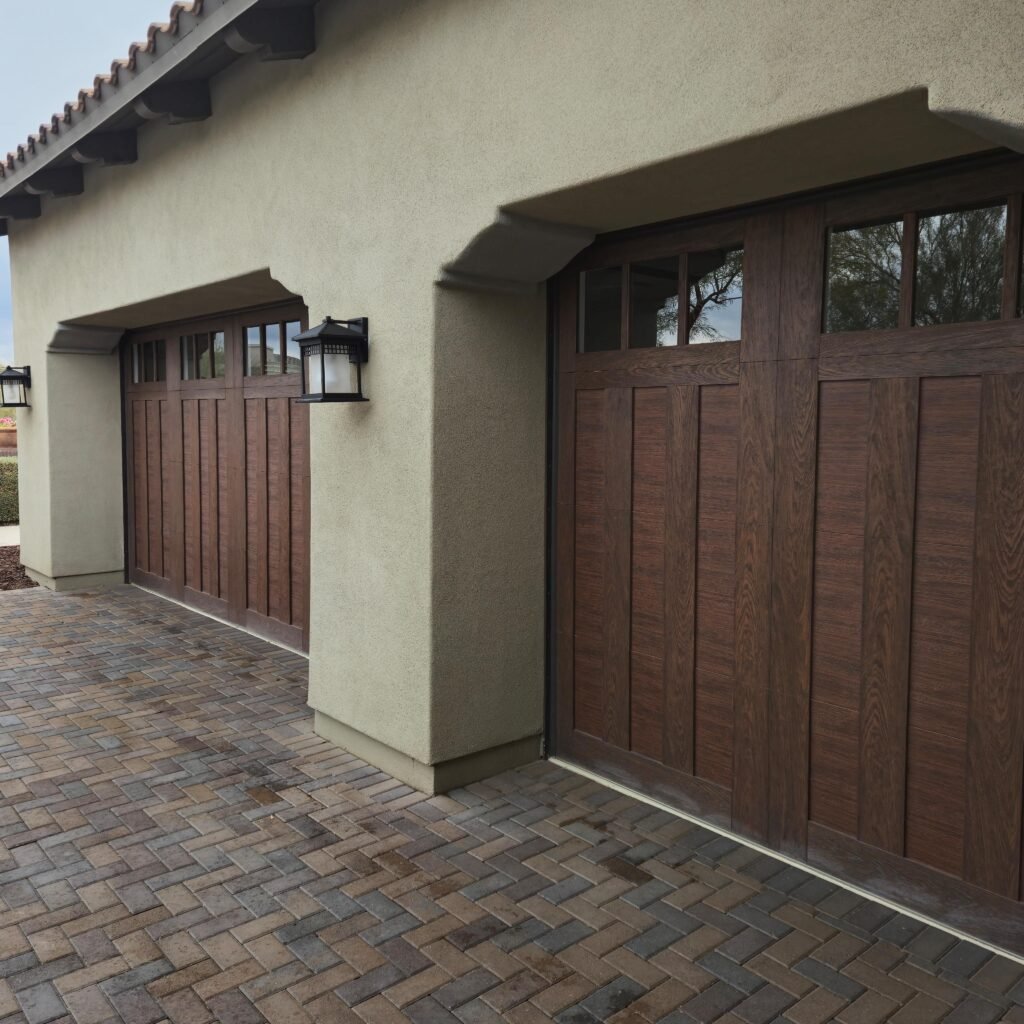 Modern home with two brown wooden garage doors featuring windows, beige stucco walls, and brick paver driveway