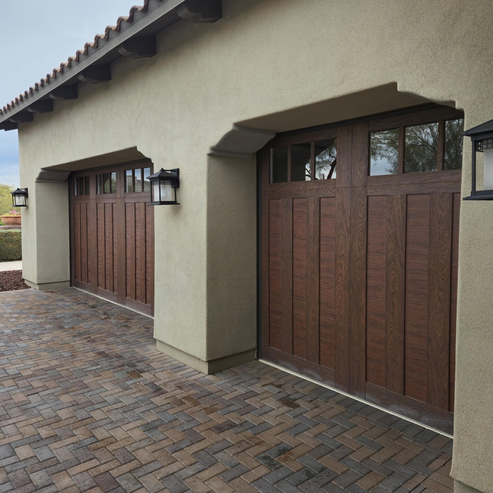 Modern home with two brown wooden garage doors featuring windows, beige stucco walls, and brick paver driveway